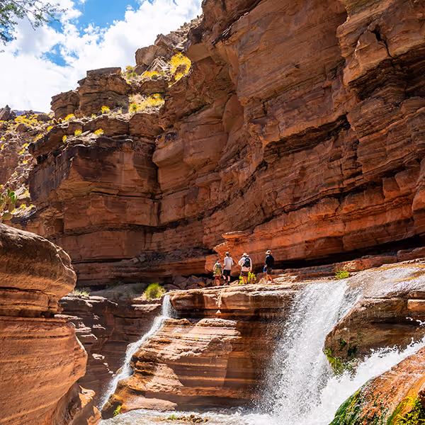 Group of hikers standing on rock ledge near waterfall inside a red rock canyon under a blue sky.